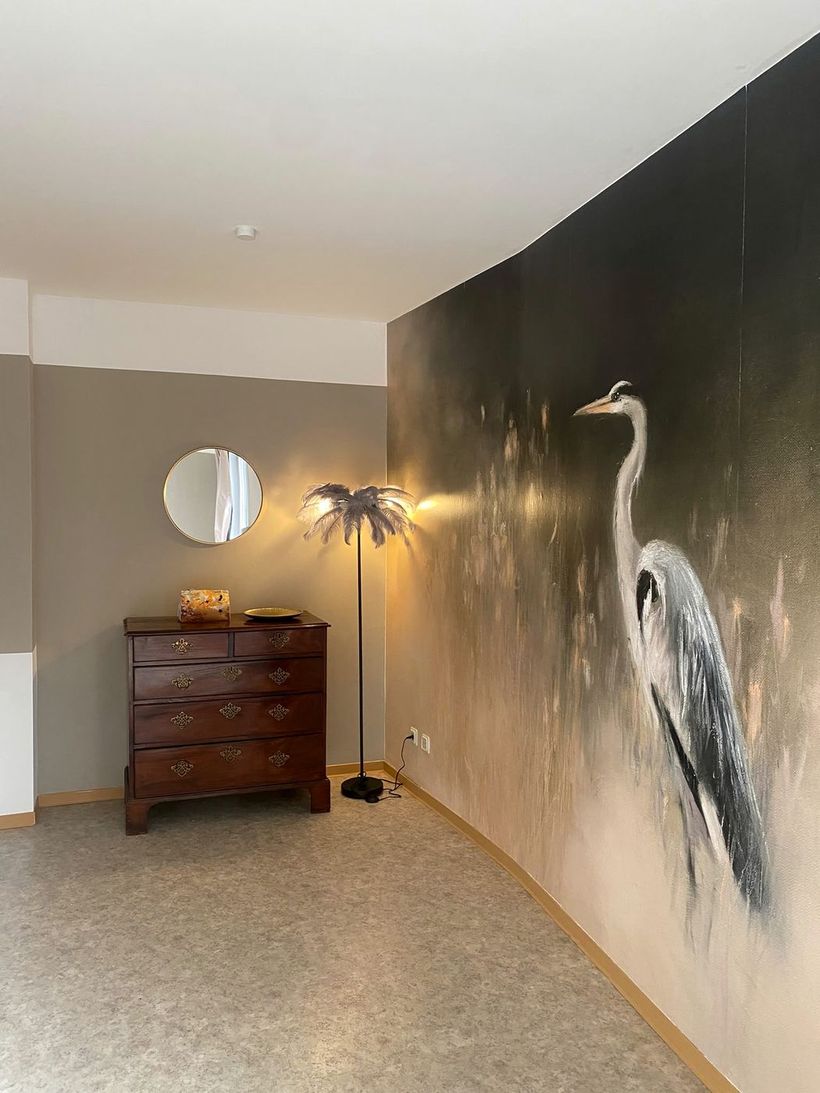 Behandlungsraum mit großem, gemaltem Reiher an der Wand, Holzkommode, rundem Spiegel und Federleuchte